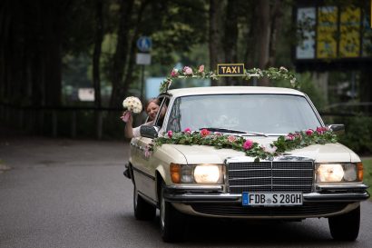 Braut winkt aus ihrem Hochzeitsauto mit ihrem Brautstrauß